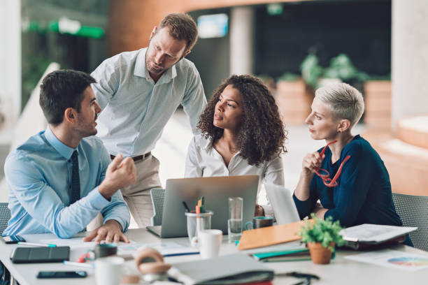 Group of diverse professionals collaborating in a modern office setting, discussing ideas around a table with laptops, documents, and coffee cups.