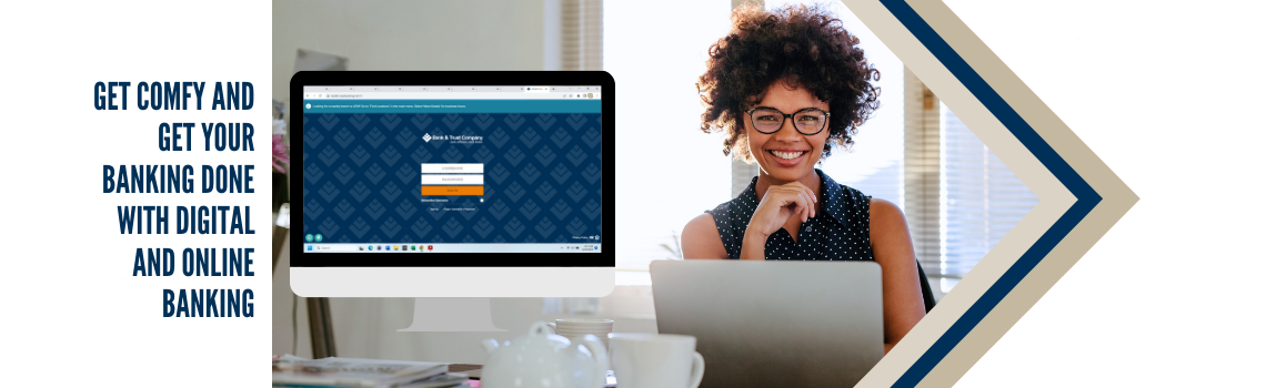 "A cheerful woman with curly hair and glasses sitting at a desk with a laptop. A computer screen displays the Bank & Trust Company online banking login page. Text on the left reads 'Get comfy and get your banking done with digital and online banking.'"